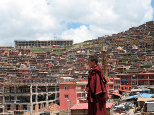 Mönch im Vordergrund, läuft vor Behausungen in Larung Gar im Hintergrund, schaut nachdenklich
