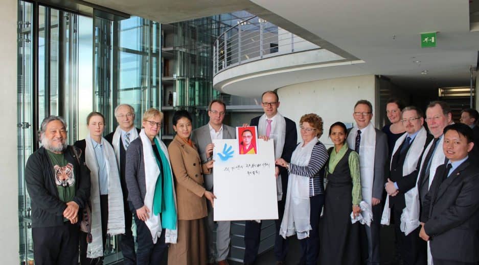 Viele Politiker und Nhyima Lhamo stehen im Halbkreis, halten ein Schild mit inem Bild von Tenzin Rinpoche in den Händen
