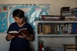 Tashi Wangchuk lehnt an einer Wand und liest in einem Buch, neben ihm ein Bücherregal